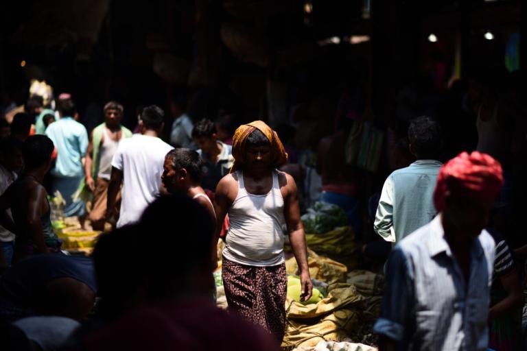A crowded market scene with people walking and standing among various stalls in dim lighting. A man in the center wears a white tank top and a towel on his head.