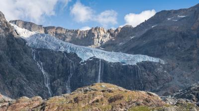 Glacier with ice cascading down between rocky peaks under a clear blue sky, flanked by waterfalls and rugged terrain.