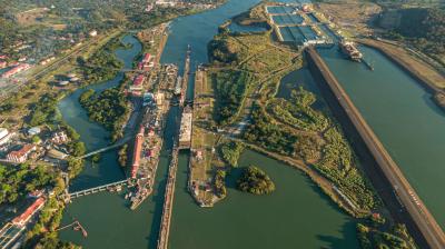 Aerial view of the panama canal with multiple lock gates open, connecting large bodies of water, surrounded by lush greenery and infrastructure.